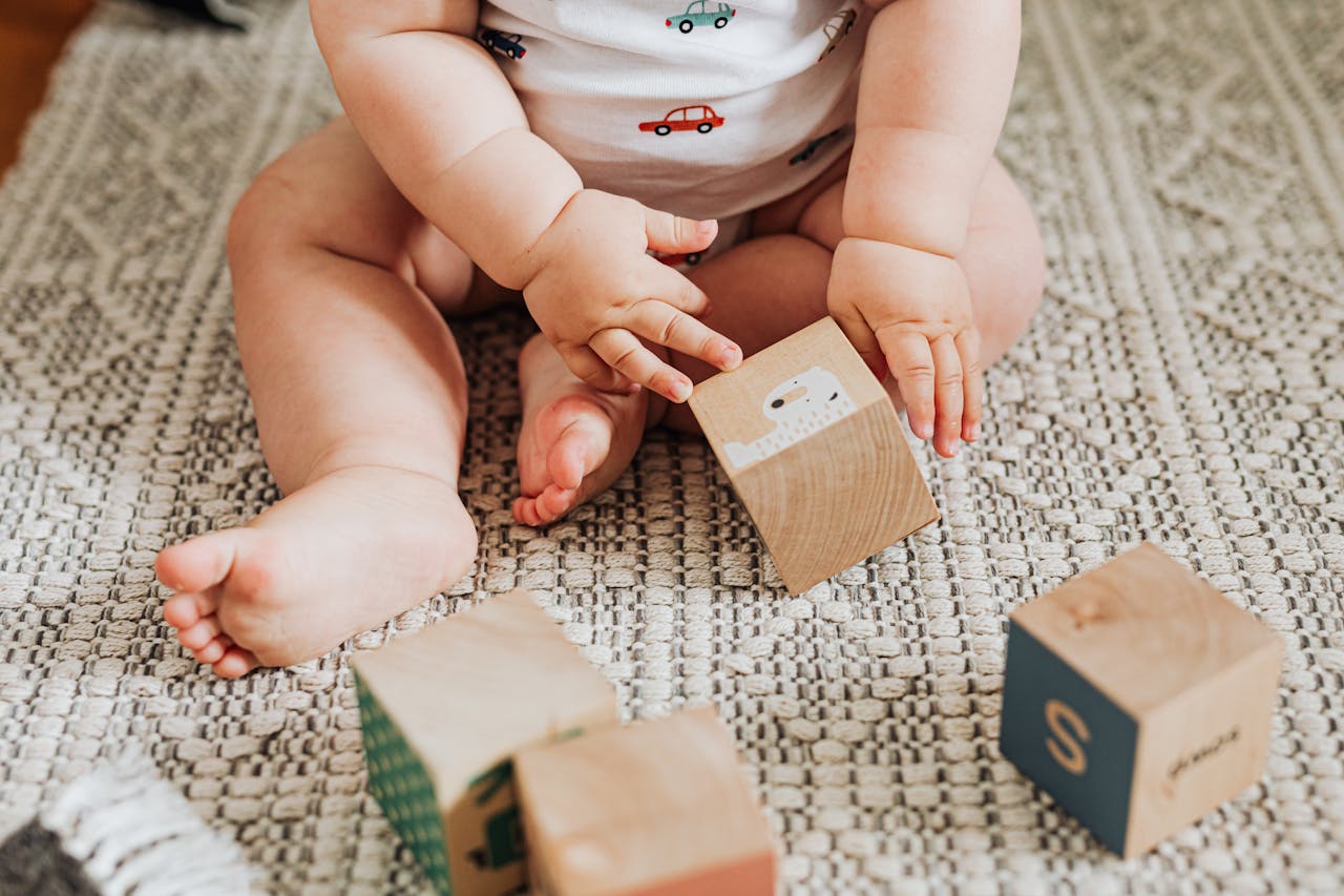 about-us-01 A baby sitting and playing with wooden toy blocks on a textured carpet indoors.